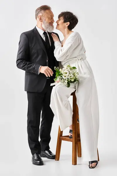 A middle-aged bride and groom, dressed in wedding gowns, sit together on a chair, celebrating their special day in a studio setting. — Stock Photo