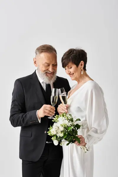 Наречена середнього віку і наречений витончено стоять поруч, тримаючи шампанські окуляри в студійній обстановці, святкуючи свій особливий день. — стокове фото