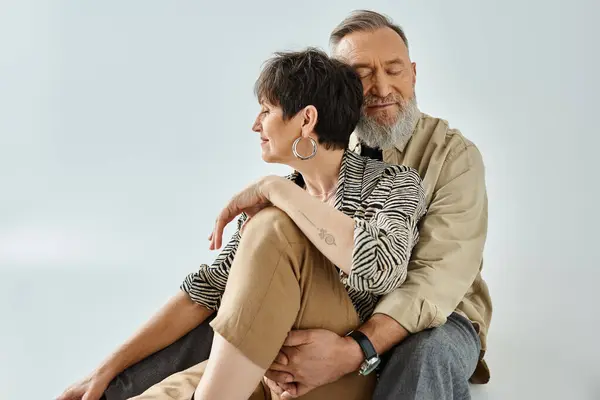 Un hombre y una mujer de mediana edad con un atuendo elegante se sientan entrelazados, creando una composición visualmente sorprendente en un entorno de estudio. - foto de stock