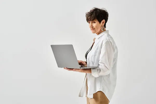 Stylish middle-aged woman in white shirt holding a laptop in a studio setting. — Stock Photo