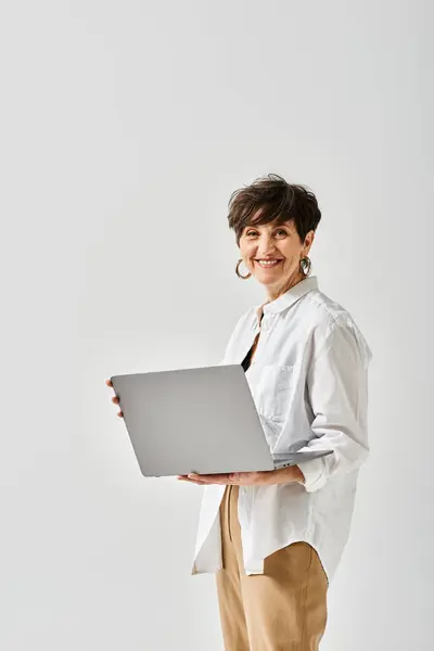 Una mujer de mediana edad con un atuendo elegante y cabello corto sostiene una computadora portátil en sus manos en un entorno de estudio. - foto de stock