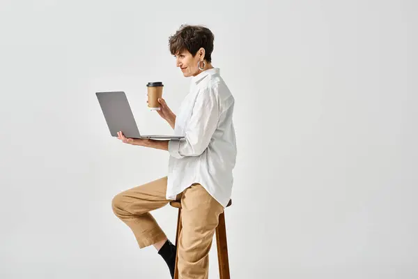 A middle-aged woman in stylish attire sits on a stool, working on a laptop in a studio setting. — Stock Photo