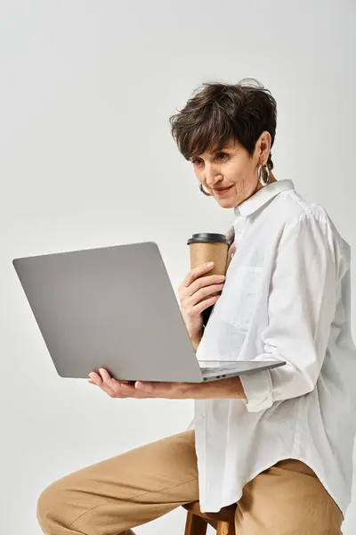 Una donna di mezza età elegantemente vestita con i capelli corti siede su uno sgabello, multitasking con una tazza di caffè in una mano e un computer portatile nell'altra. — Foto stock