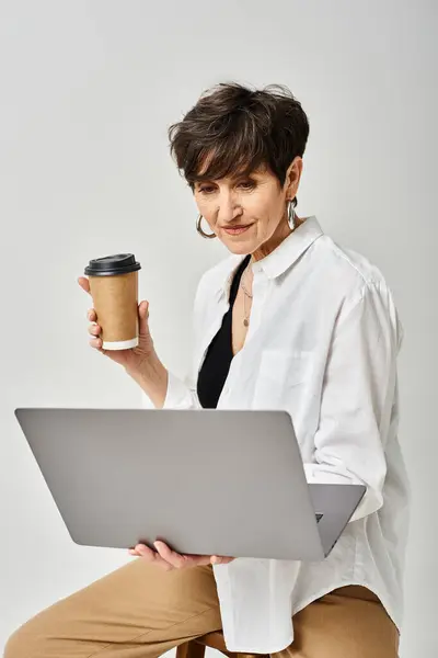 Middle aged woman multitasking, holding a coffee cup and laptop in a stylish studio setting. — Stock Photo