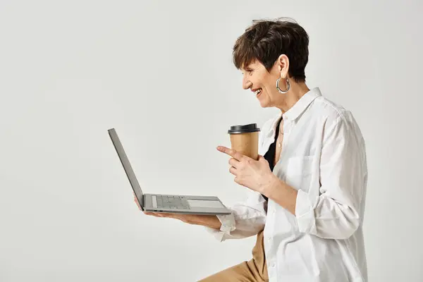 A middle aged woman in stylish attire holding a cup of coffee while working on a laptop in a studio setting. — Stock Photo