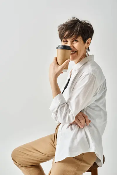 Una mujer de mediana edad elegante con el pelo corto se sienta en una silla, sosteniendo pacíficamente una taza de café. - foto de stock