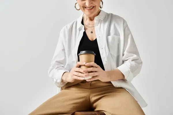 Eine Frau mittleren Alters in eleganter Kleidung sitzt auf einem Hocker und hält gelassen eine Tasse Kaffee. — Stockfoto