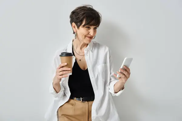 Stylish middle-aged woman with short hair multitasking, holding a cup of coffee and checking her cell phone. — Stock Photo