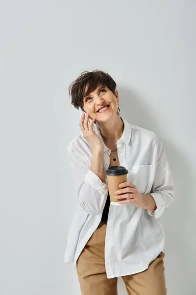 Uma mulher de meia-idade com cabelo curto, vestida elegantemente, multitarefa, segurando uma xícara de café e conversando em seu celular. — Fotografia de Stock