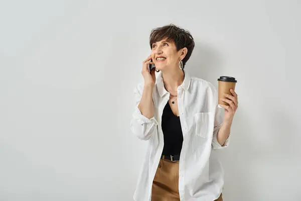 Elegante mujer de mediana edad con pelo corto multitarea, sosteniendo una taza de café mientras habla en un teléfono celular. - foto de stock