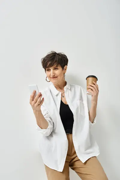 A stylish middle-aged woman with short hair is holding a cup of coffee in one hand and a cell phone in the other. — Stock Photo