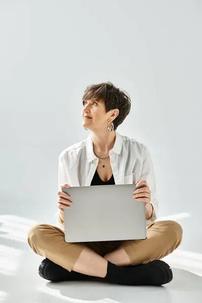 Eine Frau mittleren Alters mit kurzen Haaren, stilvoll gekleidet, sitzt auf dem Boden und arbeitet im Studio an einem Laptop.. — Stockfoto