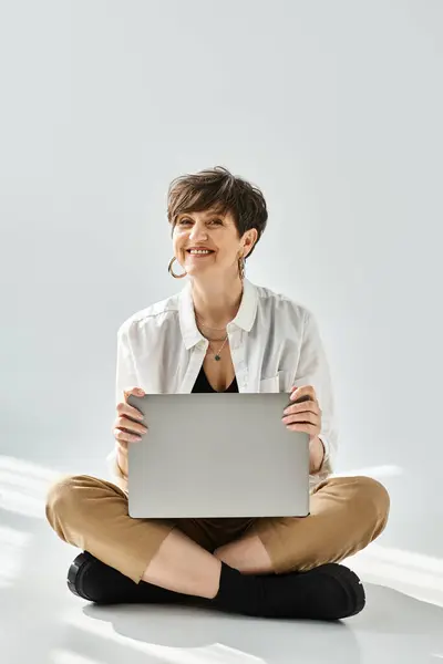 A stylishly dressed middle-aged woman with short hair is sitting on the floor, working on a laptop in a studio setting. — Stock Photo