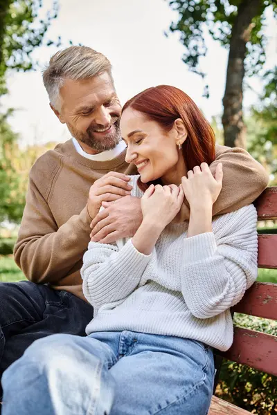A loving couple dressed casually, sitting together on a wooden bench in a serene park setting. — Stock Photo