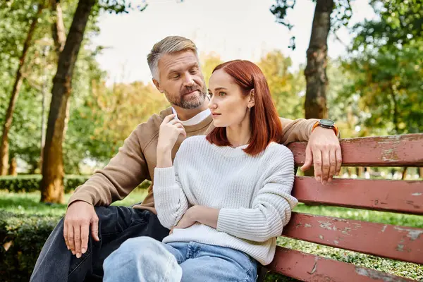 Un hombre y una mujer en atuendo casual se sientan tranquilamente juntos en un banco del parque. - foto de stock