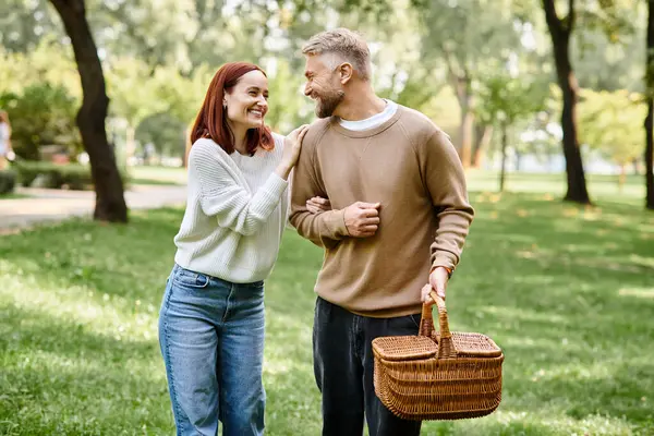 Adult couple in casual attire walking through park holding a basket. — Stock Photo