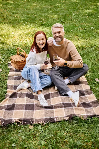 Una pareja sentada en una manta, sosteniendo copas de vino en un parque. - foto de stock