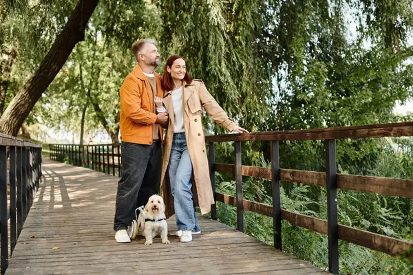 Una pareja de adultos en atuendo casual, acompañados por su perro, de pie en un puente en un parque. - foto de stock
