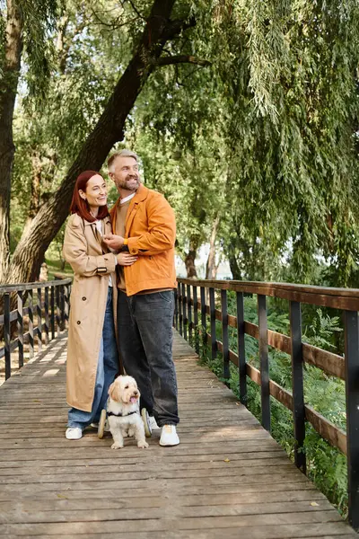 Un hombre y una mujer con un perro de pie en un puente, disfrutando de un paseo pausado. - foto de stock