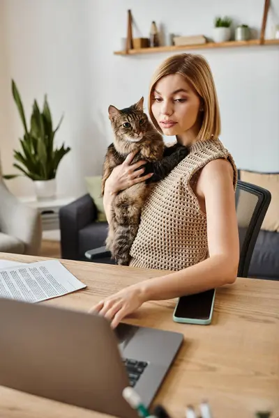 Una mujer elegante con el pelo corto sentado en una mesa de trabajo en un ordenador portátil con su gato a su lado. - foto de stock