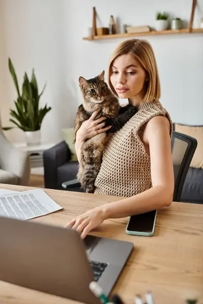 A stylish woman with short hair sitting at a table working on a laptop with her cat by her side. — Stock Photo