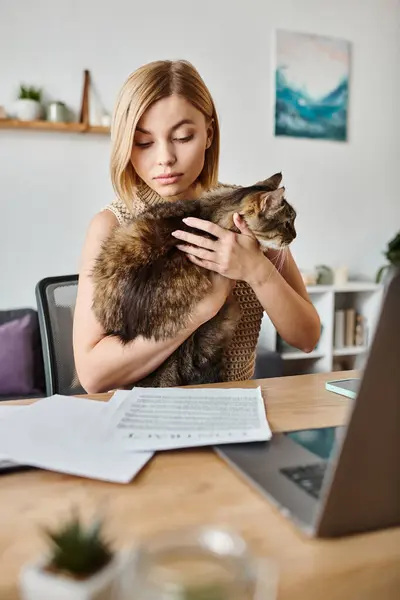 Una mujer con el pelo corto sentado en un escritorio, sosteniendo a un gato en sus brazos mientras está en casa. - foto de stock