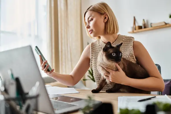 Una donna con i capelli corti tiene un gatto mentre si concentra su uno schermo del computer portatile a casa. — Foto stock