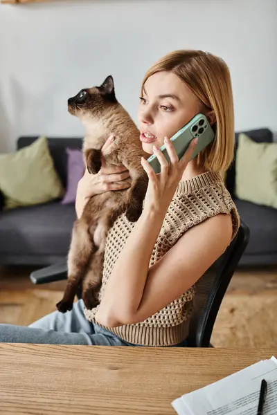 Una donna si impegna interattivamente in una conversazione telefonica mentre tiene affettuosamente il suo gatto a un tavolo. — Foto stock