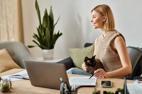 Uma mulher com cabelo curto senta-se em uma mesa com um laptop, digitando, como seu gato olha com curiosidade. — Fotografia de Stock