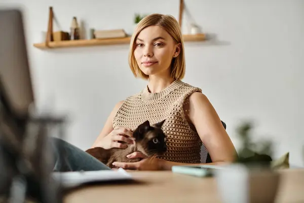A serene woman with short hair sits at a desk, tenderly holding her cat in her arms at home. — Stock Photo