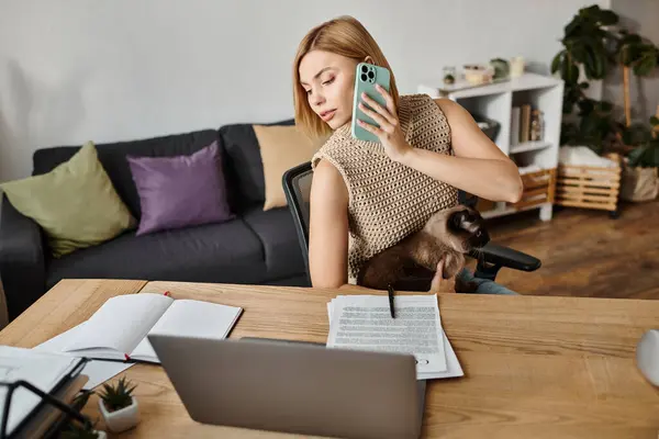 Стильная женщина с короткими волосами сидит за столом, поглощенная своим мобильным телефоном, в то время как ее кот бездельничает рядом с ней. — стоковое фото