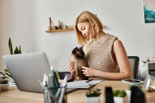 Una mujer elegante con el pelo corto sentado en un escritorio, suavemente sosteniendo a un gato en sus brazos mientras que mira calma y contenido. - foto de stock