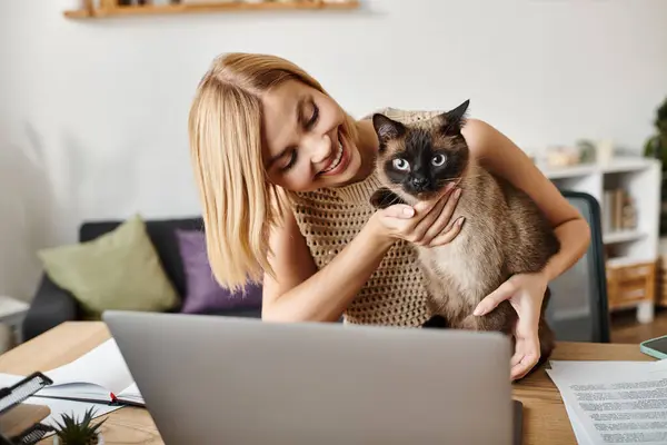 Una donna con i capelli corti tiene il suo gatto mentre guarda uno schermo del computer portatile in un ambiente accogliente casa. — Foto stock