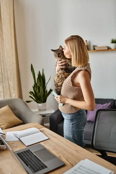Une femme sereine aux cheveux courts tient son chat, debout devant un ordinateur portable dans un cadre confortable à la maison. — Photo de stock