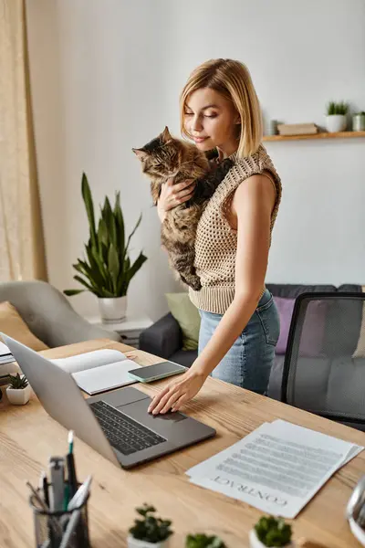 Une femme aux cheveux courts câline son chat tout en utilisant un ordinateur portable dans un cadre confortable à la maison. — Photo de stock