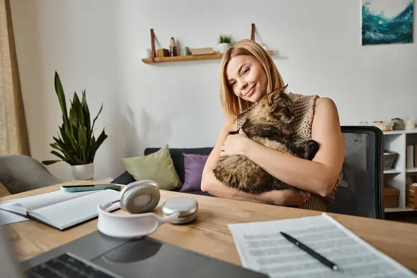 Uma mulher com cabelo curto sentada em uma mesa, segurando suavemente seu gato em um momento pacífico e amoroso em casa. — Fotografia de Stock