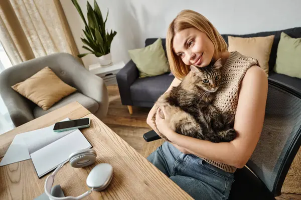 Eine Frau mit kurzen Haaren sitzt an einem Tisch, hält und streichelt ihre Katze in einem friedlichen Moment zu Hause. — Stockfoto