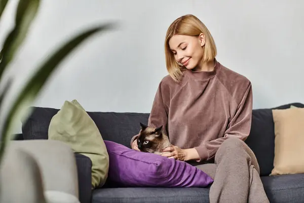 Una mujer serena con el pelo corto se relaja en un sofá, sujetando suavemente a su amado gato cerca de su pecho. - foto de stock