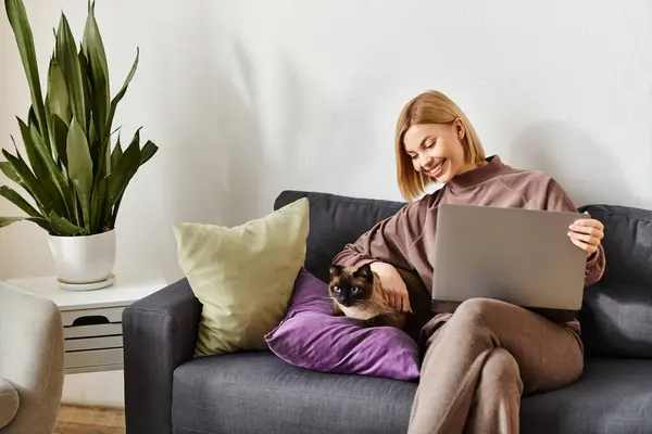 Una donna con i capelli corti si rilassa su un divano con il suo gatto al suo fianco, lavorando su un computer portatile in un ambiente accogliente casa. — Foto stock