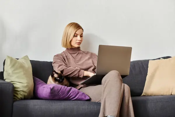 A stylish woman with short hair focusing on her laptop while relaxing on a comfortable couch, with her cat by her side. — Stock Photo