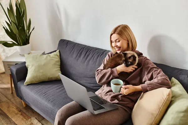Una donna con i capelli corti seduta su un divano, coccolare un gatto e lavorare su un computer portatile in un ambiente accogliente casa. — Foto stock