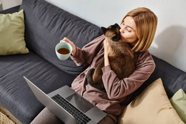 Una mujer atractiva con el pelo corto sentado en un sofá, sosteniendo un gato en sus brazos y una taza de café en la mano. - foto de stock