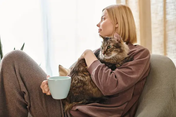 Una mujer serena con el pelo corto se sienta en un sofá sosteniendo una taza de café y un gato amistoso en un ambiente acogedor en casa. - foto de stock