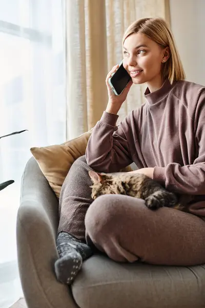 Une femme élégante avec des cheveux courts bavardant sur son téléphone portable, partageant un moment avec son adorable chat sur le canapé. — Photo de stock