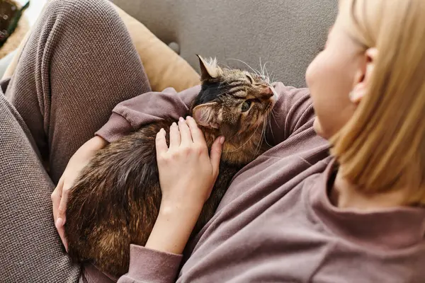 Una mujer con el pelo corto reclinado en un sofá, acariciando tiernamente a su gato con una expresión serena. - foto de stock