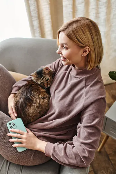 A stylish woman with short hair relaxes in a chair while gently holding her beloved cat. — Stock Photo