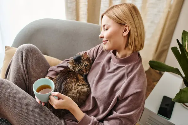 Стильная женщина отдыхает на диване, наслаждаясь чаем и обнимая содержательную кошку в спокойной домашней обстановке. — стоковое фото