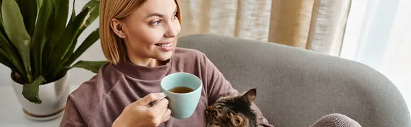 A woman with short hair sits on a couch, holding a cup of coffee and a cat in her arms, enjoying a cozy moment at home. — Stock Photo