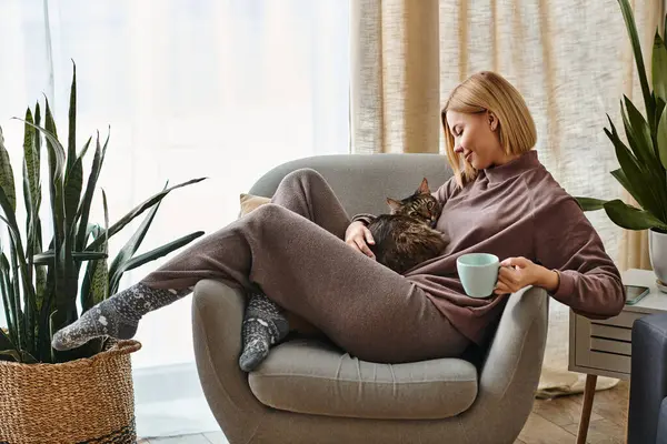 Eine Frau mit kurzen Haaren entspannt sich auf einem Stuhl, während sich ihre Katze auf ihrem Schoß niederlässt. — Stockfoto