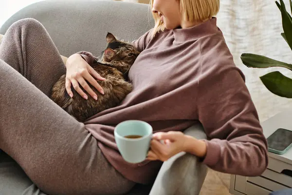 A woman with short hair relaxes on a couch, cradling a cup of coffee and a content cat in her arms. — Stock Photo